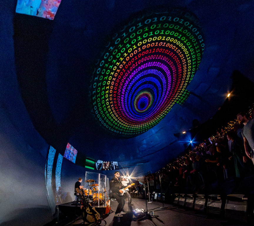 A band performs on stage with colorful light effects and a large visual projection.