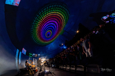 A band performs on stage with colorful light effects and a large visual projection.