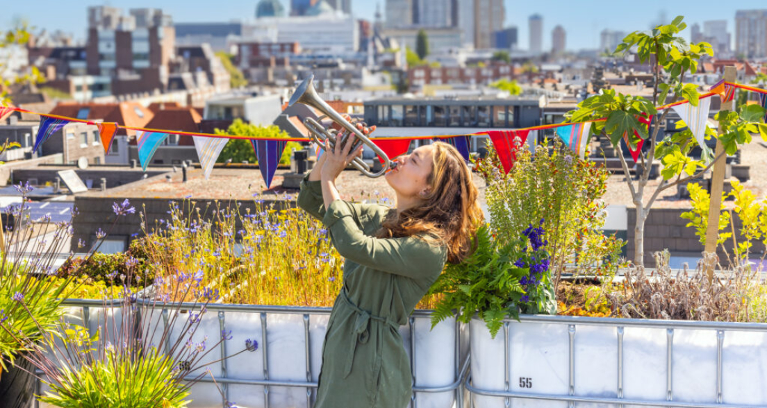 A woman in a green coat plays trumpet on a rooftop.