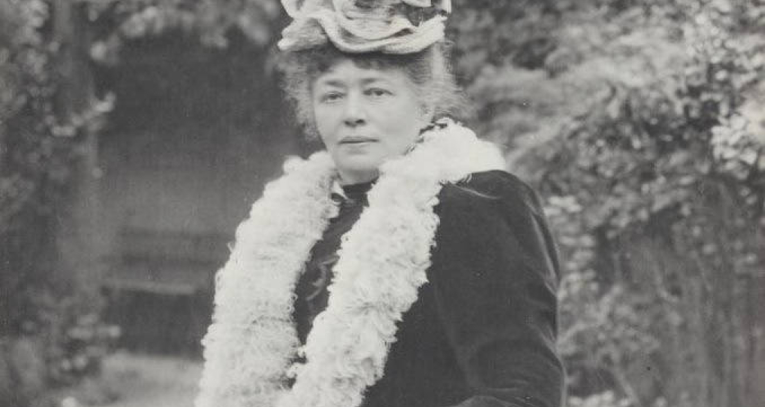 A woman with a unique hat and fur collar poses in a green garden.