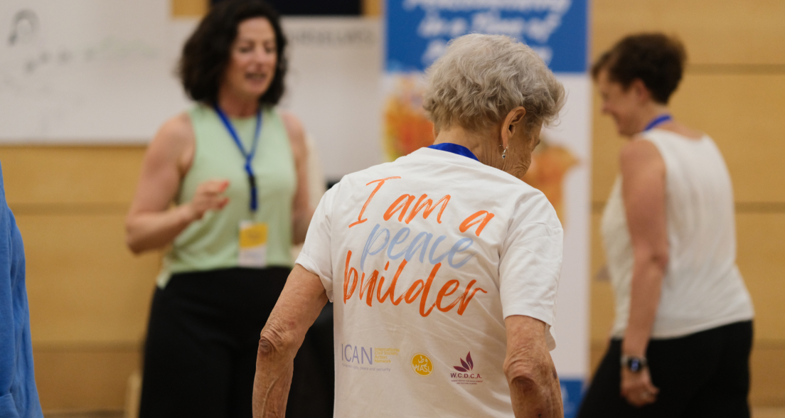 An elderly woman in a T-shirt with 'I am a peace builder' in a dance class.