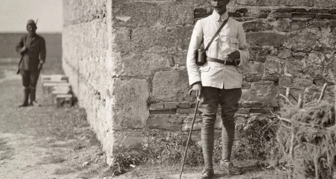 Major Roelfsema standing with cane and binoculars beside a stone wall at the location in Durrës, Albania, where Major Thomson was fatally shot during the 1914 peace mission. An armed soldier stands guard in the background.