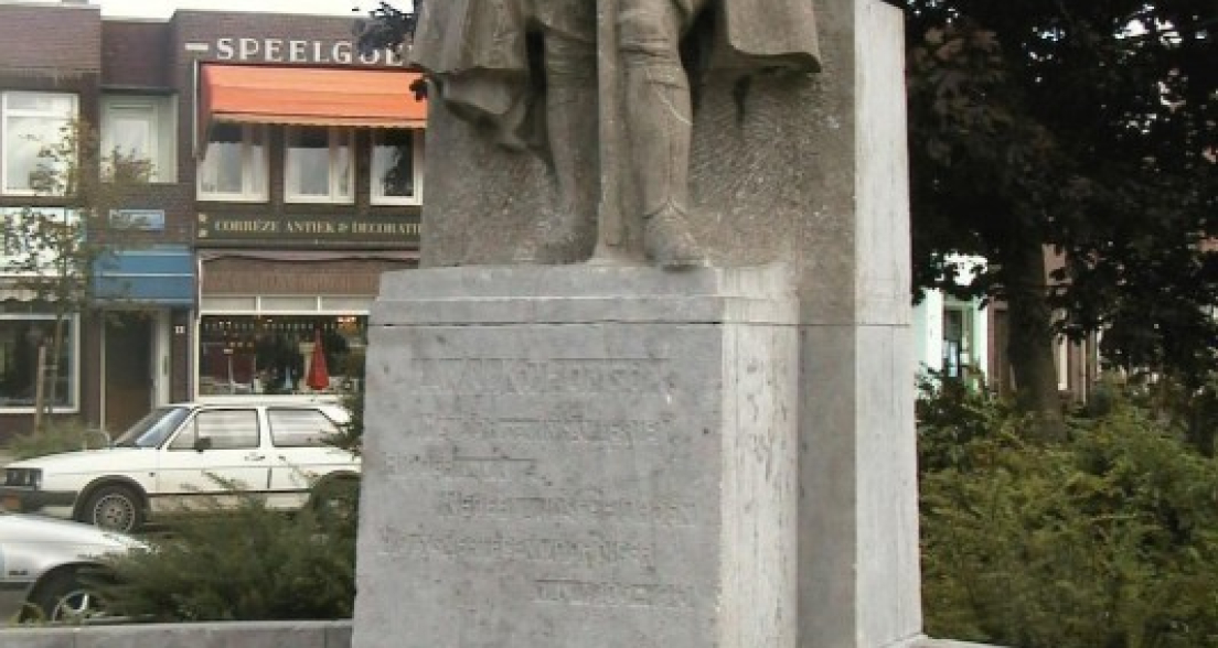 Stone statue of Major L.W.J.K. Thomson in military uniform and cape, standing atop a pedestal at Thomsonplein in The Hague, flanked by a semicircular stone bench and set against a backdrop of local shops.