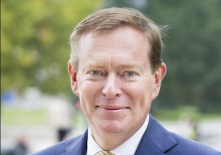 A man in a blue blazer and yellow tie smiles in a park.