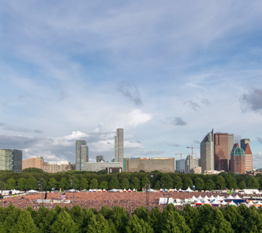 Een groot publiek bij een buitenconcert in Den Haag, met hoge gebouwen op de achtergrond.