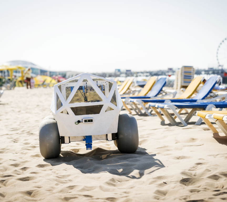 A robot is positioned on the sandy beach with deck chairs in the background.