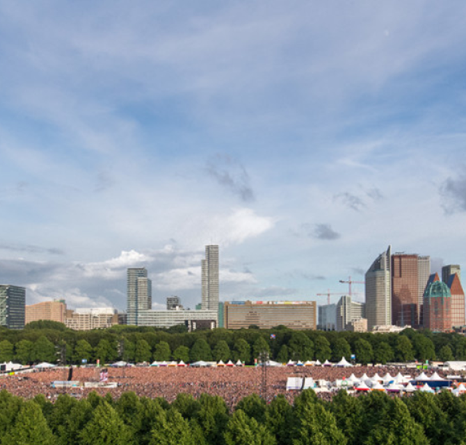 Een groot publiek bij een buitenconcert in Den Haag, met hoge gebouwen op de achtergrond.