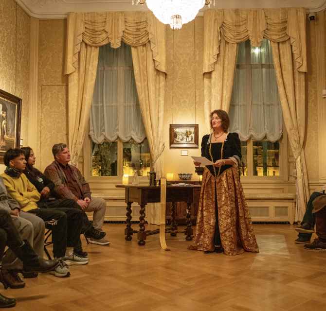 A woman in historical clothing presents to an audience in a hall.