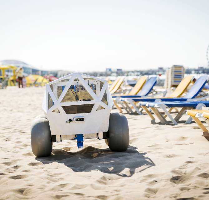 A robot is positioned on the sandy beach with deck chairs in the background.
