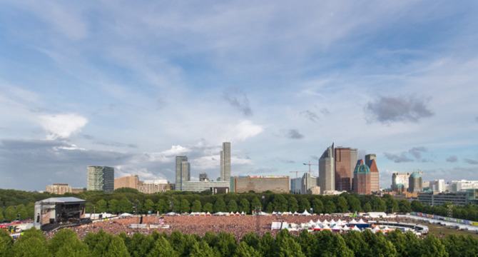 Een groot publiek bij een buitenconcert in Den Haag, met hoge gebouwen op de achtergrond.