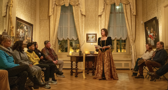A woman in historical clothing presents to an audience in a hall.