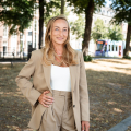 A woman with long, blonde hair is wearing a beige blazer and pants, standing in a park.