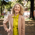 Woman with curly hair in a green blouse and pink blazer, smiling in a park