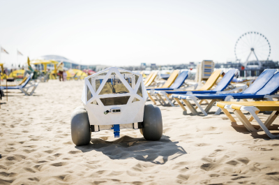 A robot is positioned on the sandy beach with deck chairs in the background.