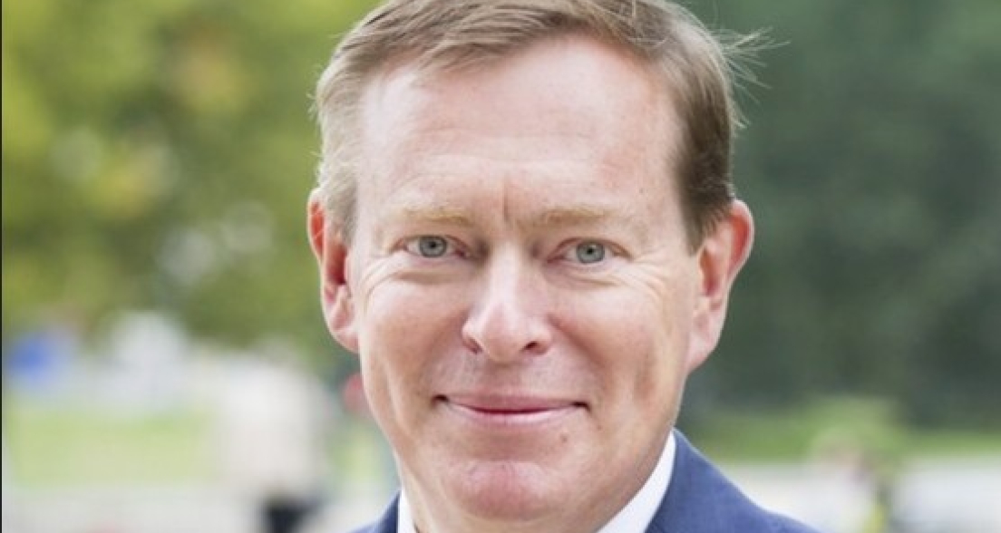 A man in a blue blazer and yellow tie smiles in a park.