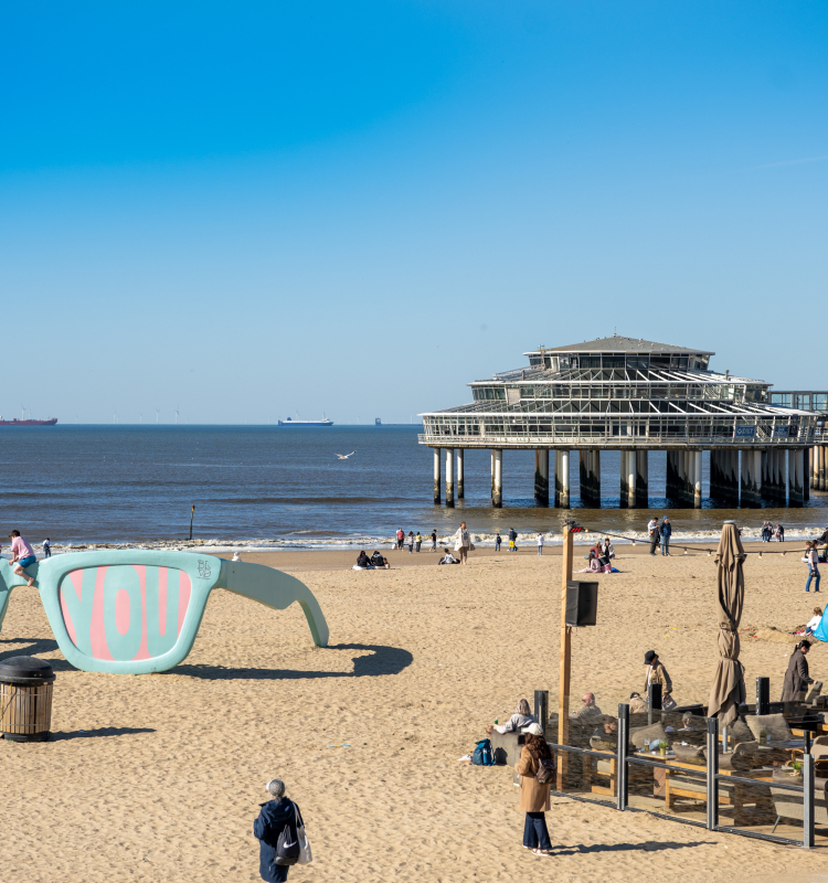 People on the beach at Scheveningen Pier with large sunglasses and clear blue sky.